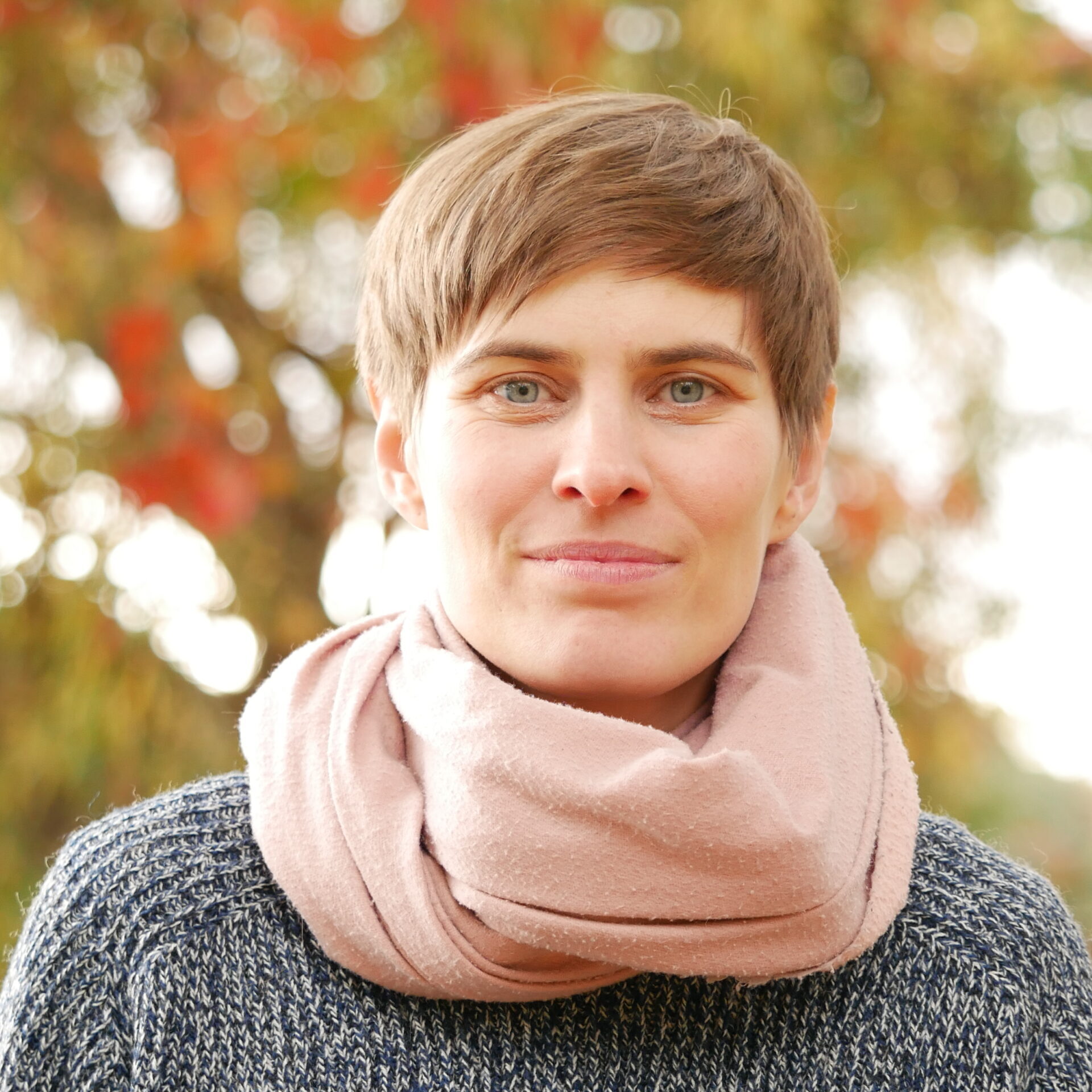 Foto von Anne, schaut zugewandt in die Kamera. Im Hintergrund ein herbstlicher Baum.