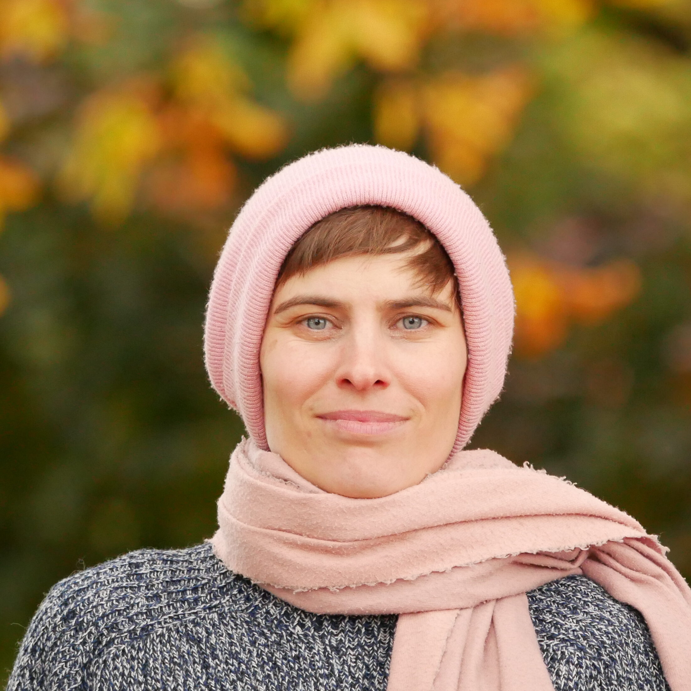 Anne in rosé-farbenem Schal und Mütze, schaut freundlich in die Kamera. Im Hintergrund ein herbstlicher Baum.